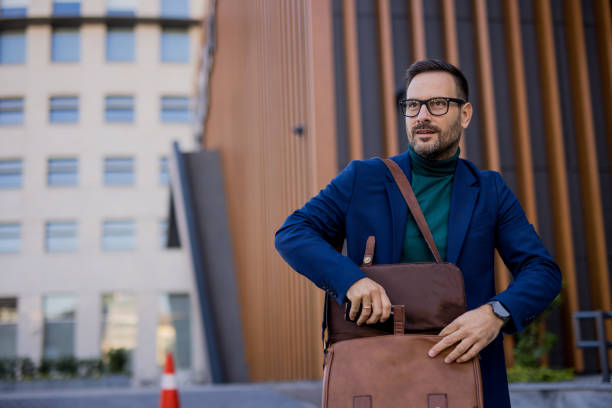 istock handsome and attractive man is on his way to work, walking down the street while taking his laptop out of his bag. In the background is a modern building, and he is elegantly dressed in a dark blue suit, radiating confidence and style. 2180223694