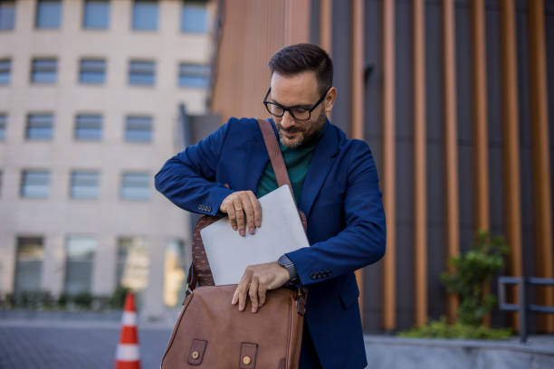 istock handsome and attractive man is on his way to work, walking down the street while taking his laptop out of his bag. In the background is a modern building, and he is elegantly dressed in a dark blue suit, radiating confidence and style. 2180223697