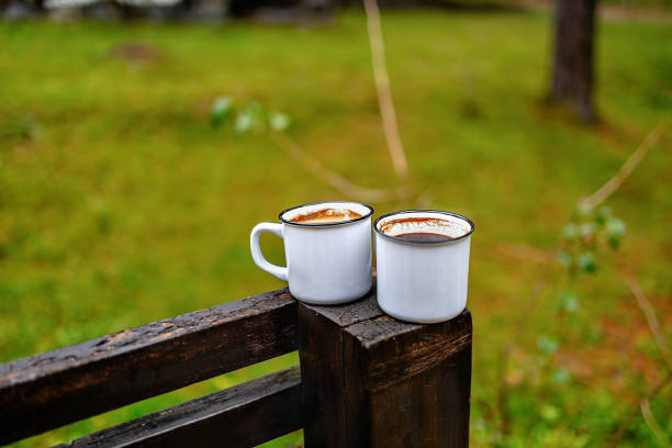 Two white mugs filled with coffee rest on a wooden railing, with a lush green background enhancing the tranquil atmosphere of morning reflection.