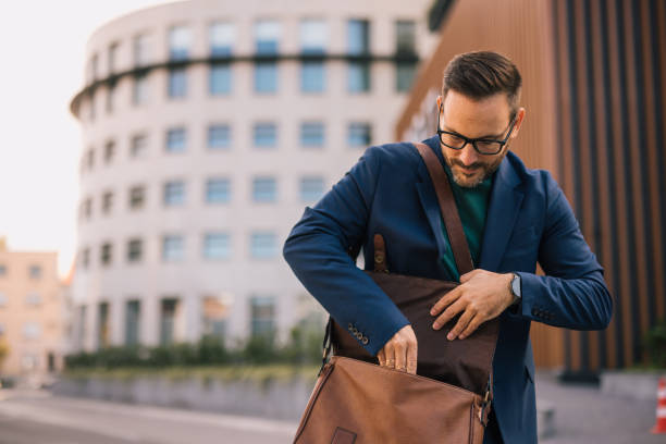 istock handsome and attractive man is on his way to work, walking down the street while taking his laptop out of his bag. In the background is a modern building, and he is elegantly dressed in a dark blue suit, radiating confidence and style. 2180224888