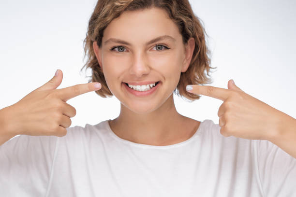 istock Smiling Young Woman Pointing at Her White Teeth on White Background 2180231475