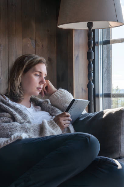 istock A Woman Reading on Her E-Reader book Quietly by a Large Window in a Cozy, Rustic Living Room 2180238648