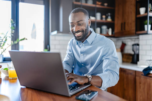 istock A smiling African-American man is sitting at a kitchen counter using a laptop. He is wearing a light blue shirt and is engaged with the screen, possibly working or browsing online. 2180243120