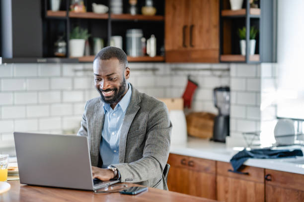 istock A smiling African-American man is sitting at a kitchen counter using a laptop. He is wearing a light blue shirt, and grey blazer and is engaged with the screen, possibly working or browsing online. 2180246281
