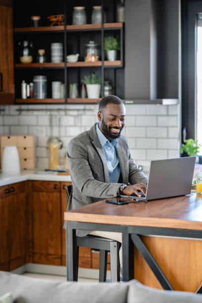 istock A smiling African-American man is sitting at a kitchen counter using a laptop. He is wearing a light blue shirt, and grey blazer and is engaged with the screen, possibly working or browsing online. 2180246533