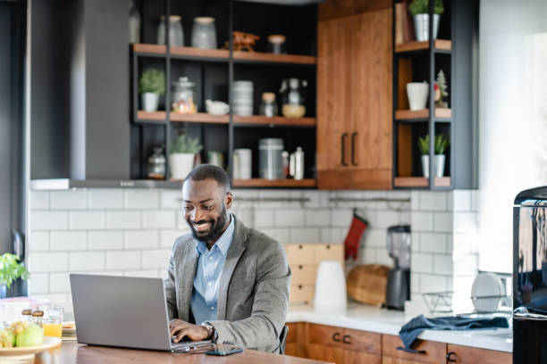 istock A smiling African-American man is sitting at a kitchen counter using a laptop. He is wearing a light blue shirt, and grey blazer and is engaged with the screen, possibly working or browsing online. 2180246703