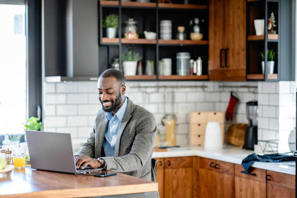 istock A smiling African-American man is sitting at a kitchen counter using a laptop. He is wearing a light blue shirt, and grey blazer and is engaged with the screen, possibly working or browsing online. 2180246705