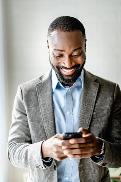 istock Smiling young man in a suit using his smartphone, looking down at the screen with focus. Professional and casual in a bright, modern setting. 2180247767