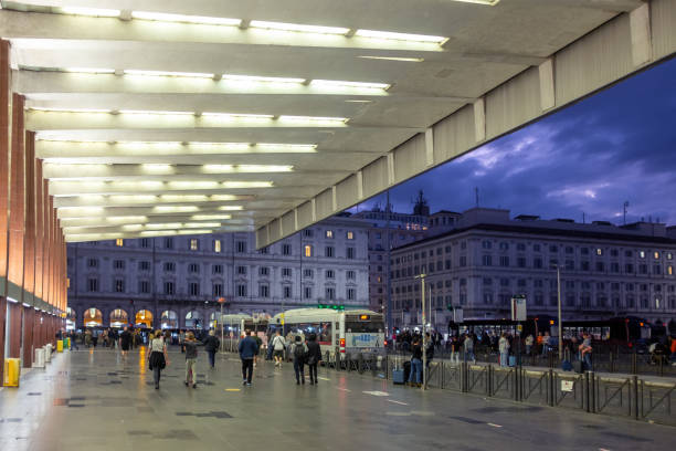 Exterior view of Roma Termini railway station building on October 21st, 2024, just after sunset, with with travellers and people walking to the surrounding streets under a large illuminated roof overhang. Roma Termini station is Rome’s largest train transportation hub.