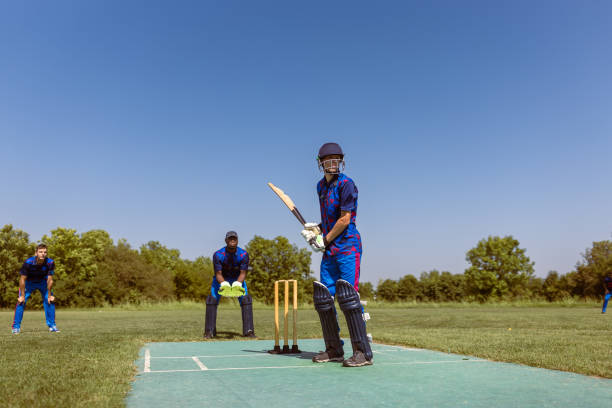 A cricket player in blue uniform is getting ready to bat while other players and the wicketkeeper stand in position on a green pitch during a sunny day.