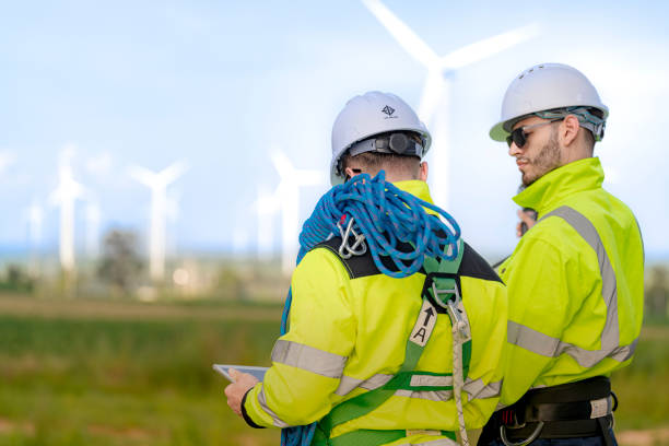 istock Two engineers in high visibility jackets and helmets are inspecting a wind farm. One is holding a mobile device, while the other is equipped with ropes for climbing, with turbines visible in the background. 2180266110