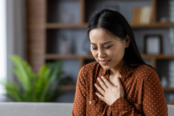 istock Concerned asian woman holding chest in thoughtful expression while seated on sofa in cozy living room interior setting. Pensive asian lady reflects emotion through serene gestures. 2180269220