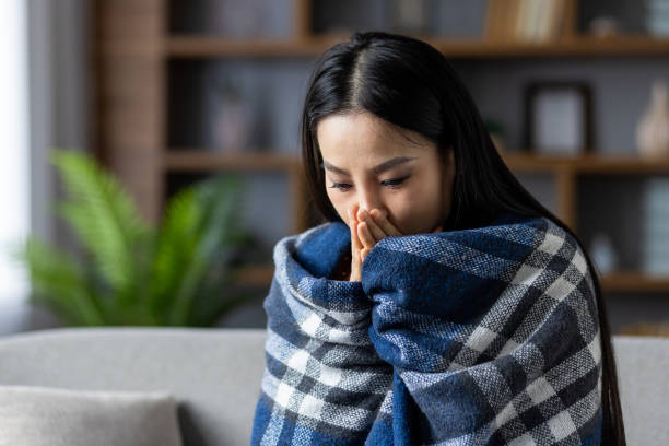istock Young woman wrapped in blue plaid blanket feeling cold inside modern apartment while sitting on cozy couch in warm home environment. Emotions of chill and comfort captured indoors. 2180272141