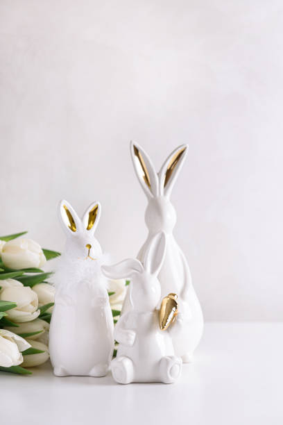 istock Three Easter rabbits figurines with bouquet of white tulips on white background. Easter celebration concept. Copy space. Front view 2180276354
