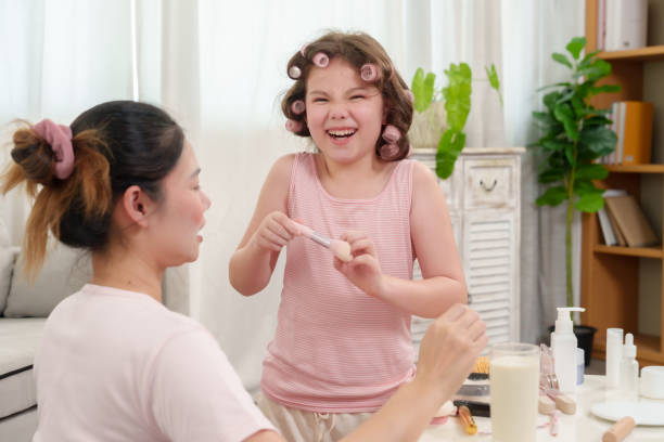 istock Young girl with curly hair rollers holding makeup brush while smiling joyfully during playful makeup session with single mother in living room. Scene filled with warmth, joy, bonding, and happiness. 2180290266