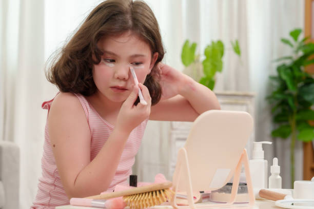 istock Young girl carefully applying makeup under her eyes while sitting at a table. The scene shows her concentration as she learns to use makeup, capturing the joy and innocence of the moment. 2180290294