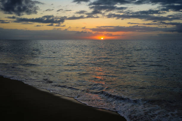 The sun dips below the horizon, casting a warm glow across the calm waters of Kalalau Beach, as the day transitions to night amidst softly rolling waves.