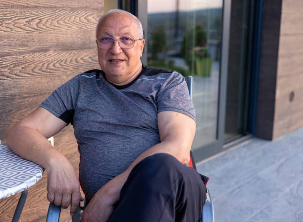 An elderly man with a warm smile sits comfortably outside on a sunny day, embracing the tranquility and peace of outdoor leisure. The scene conveys joy and contentment.