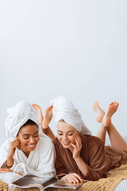 istock Two pretty ladies in bathrobes laying on bed, reading magazines 2180314267