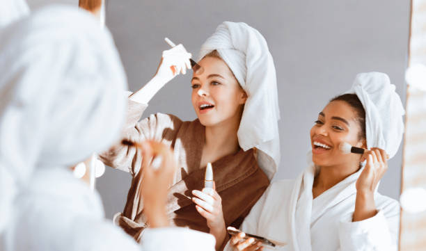 istock Cheerful girls standing in front of mirror, applying foundation 2180314331