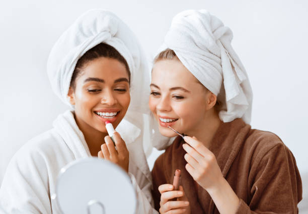 istock Joyful girlfriends in bathrobes putting lipsticks, looking at mirror 2180314400