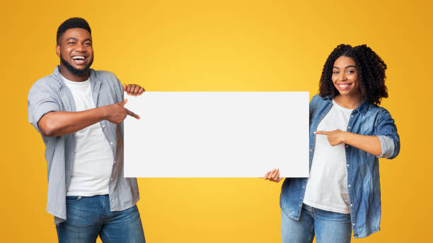 istock Cheerful black couple holding and pointing at blank advertisement placard 2180322677
