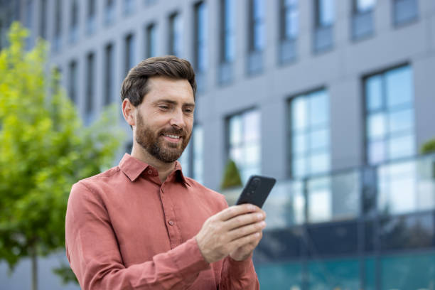 istock Smiling businessman in casual attire using smartphone outdoors in urban setting, reflecting positive communication and connectivity. Engaged in checking messages or emails during break 2180323008