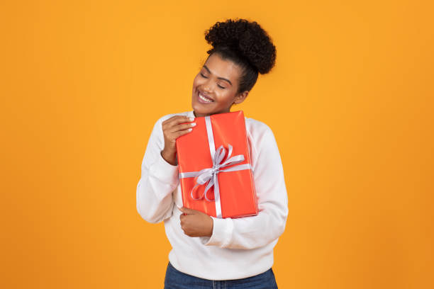 istock Young woman joyfully embracing a wrapped gift against a bright orange background 2180324453