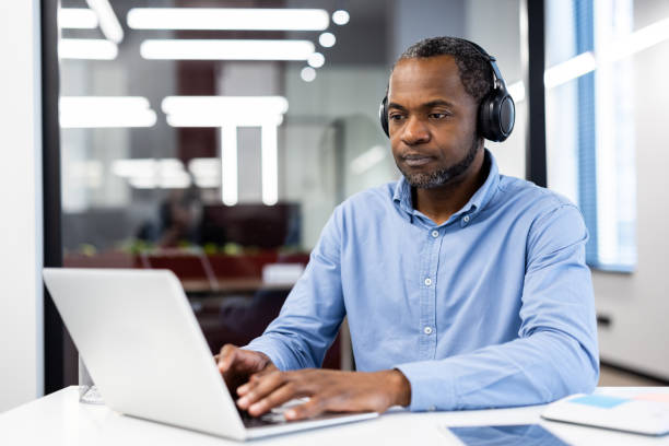 istock Focused professional man working on laptop with headphones in modern office setting, demonstrating concentration and productivity in a corporate environment. 2180325572