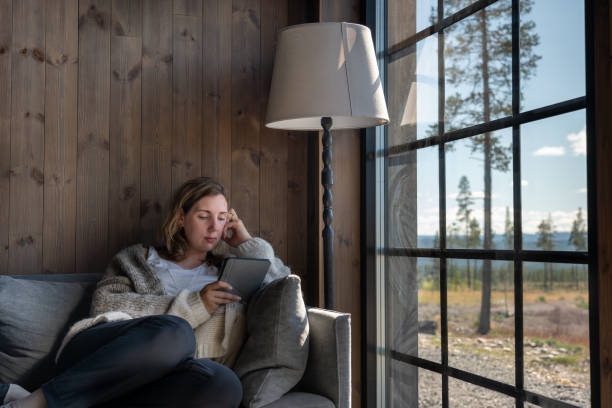Woman Reading on a E-Reader by the Window in a Cozy Rustic Cabin in the Woods During the Day