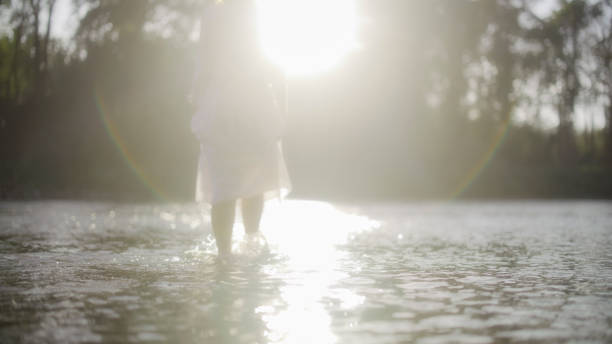 A mature Caucasian woman stands in a river, sunlight enveloping her relaxed frame, wearing light attire ideal for water, embodying tranquility in a natural setting.