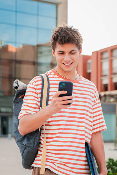istock Vertical. Male student having fun with a cellphone posting on the social media at campus. Real teenage guy smiling using a smartphone. Young man holding a mobile phone. Teen schoolboy reading a phone 2180380989
