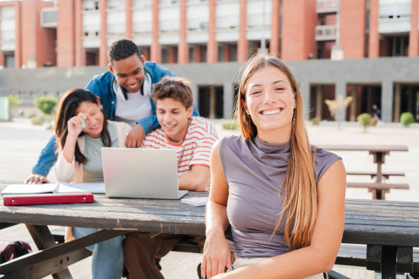 istock Young caucasian female student, smiling and looking at camera sitting with her classmates at university campus. Blonde schoolgirl staring front behind her multiracial friends while they use a laptop 2180383032