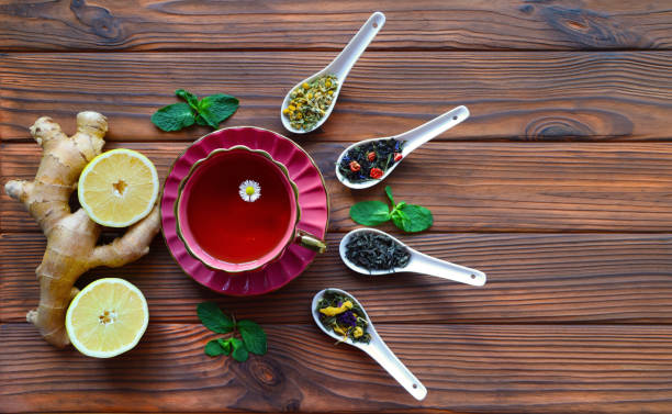 istock Cup of herbal tea with ginger, lemon, and loose leaf blends on wooden surface. A beautiful setup featuring a cup of herbal tea with a floating daisy, surrounded by ginger root, lemon halves, and four porcelain spoons filled with loose leaf tea blends. 2180444347