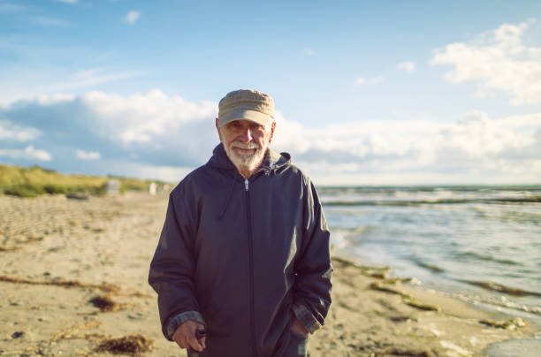 A serious looking man is looking thoughtfully in the direction of the ocean. The man has glasses, a white beard and a sailor cap on his head