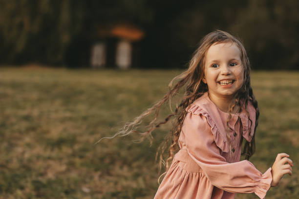 A young girl with long blonde hair raises her arms gracefully against a sunlit background. She wears a brown quilted jacket and a pink tutu, combining a cozy look with ballet elegance. The bright sunlight creates a radiant halo around her, emphasizing her poised expression and capturing a moment of joyful movement and serenity in a natural outdoor setting.