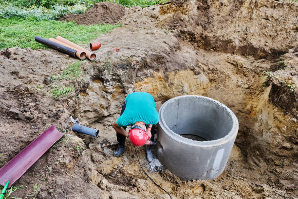 istock Worker chisels out hole for sewer pipe in concrete septic tank ring using hammer drill with chisel. 2180459480