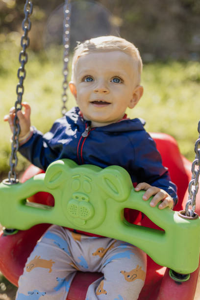 A one-year-old boy is smiling as he swings in a park on a sunny day. The scene captures the joy and simplicity of childhood in a peaceful outdoor setting