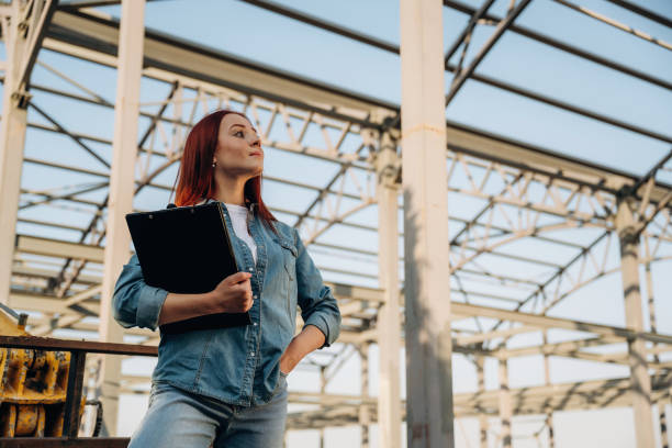 istock Holding notepad. Young redhead woman is standing near unfinished building, construction 2180470485
