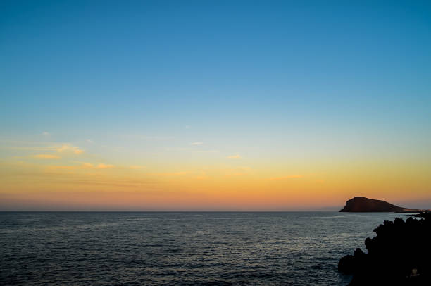 The sky is blue and the sun is setting. The water is calm and the horizon is in the distance