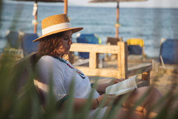 istock Young woman reading book on the beach 2180471111