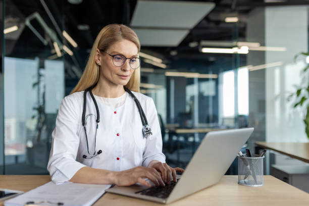 istock Confident mature doctor using laptop in modern office while wearing white coat and stethoscope 2180485784