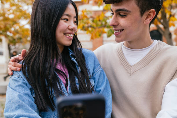 istock Young multiethnic couple smiling and looking at each other while using a smartphone outdoors 2180492510