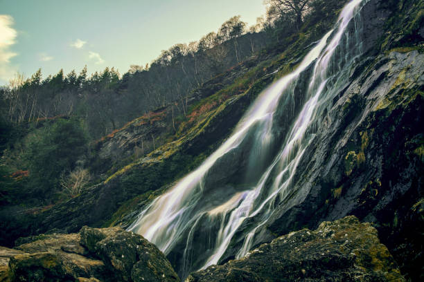 A picturesque waterfall cascades down, surrounded by lush trees and rugged rocks on the scenic hillside, creating a tranquil and serene atmosphere