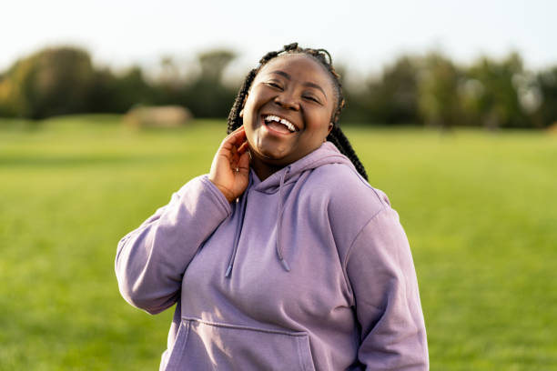 Young woman wearing a hoodie is laughing in a park Young woman laughing and running her fingers through her hair in a park, radiating happiness and confidence in her trendy style, celebrating wellbeing and self love on a sunny day plus size woman fashion laughing stock pictures, royalty-free photos & images
