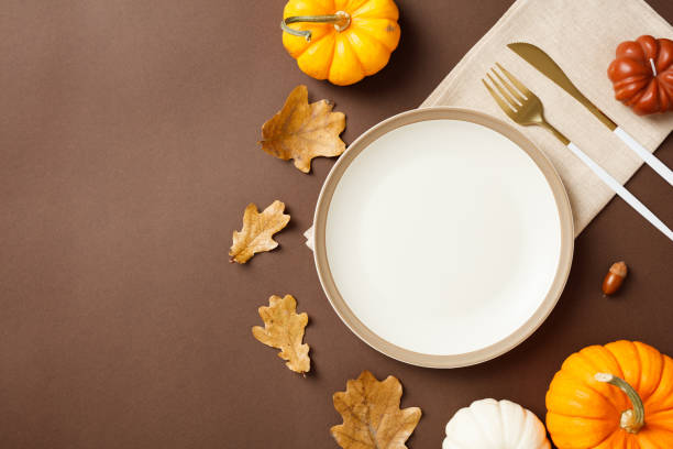 istock Autumn table setting with empty plate surrounded by colorful pumpkins, dry leaves, and cutlery on a brown background 2180502513