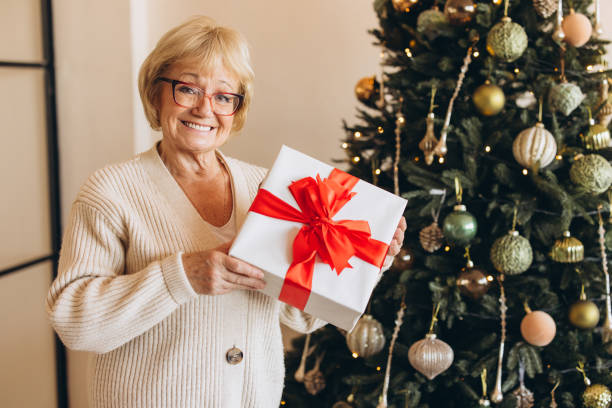 istock Smiling Senior Woman Holding Gift by Christmas Tree 2180510542