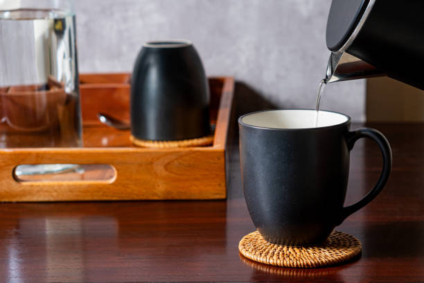 A black coffee mug placed on a woven coaster, with hot water being poured from a kettle. In the background, there is a wooden tray with a glass and another black cup. The setting is cozy