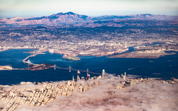 Aerial view of San Francisco Bay and the Bay Bridge with the fog rolling in over the financial district and the port of Oakland, Berkeley, etc. in the background. Mount Diablo is in the far background. John M.Lund