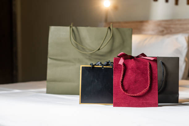 A collection of shopping bags in various sizes and colors placed on a bed. The bags include a large green bag, a medium black bag, and a small red bag with a bow. The setting is cozy and inviting